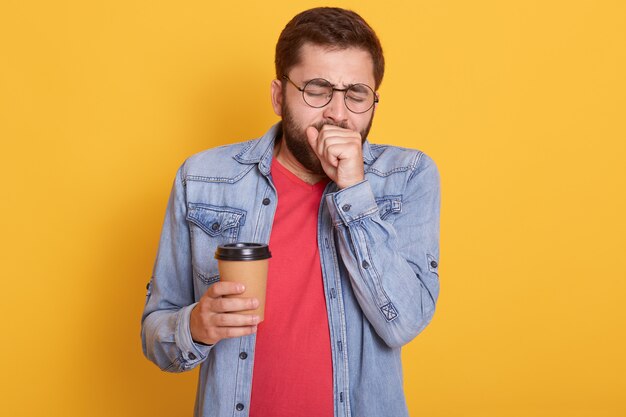 Indoor image of tired exhausted bearded guy putting hand to mouth, yawning, having desire to sleep, holding coffee in paper cup