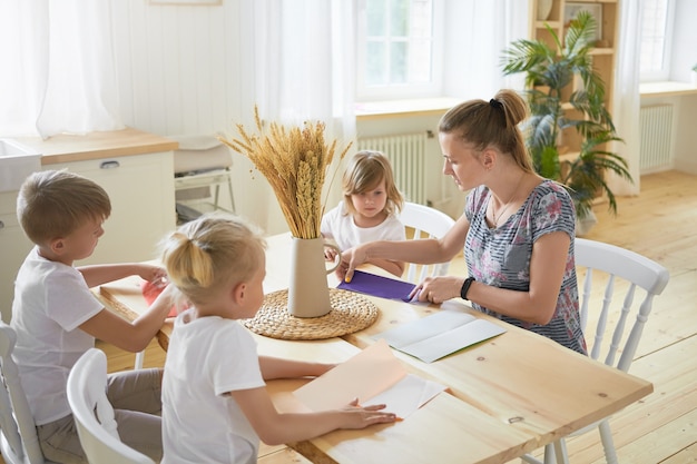 広々としたリビングルームのダイニングテーブルに座って、子供たちに折り紙の作り方を教える若い女性のベビーシッターの屋内画像。家で母親と一緒に紙飛行機を作る3人の子供。