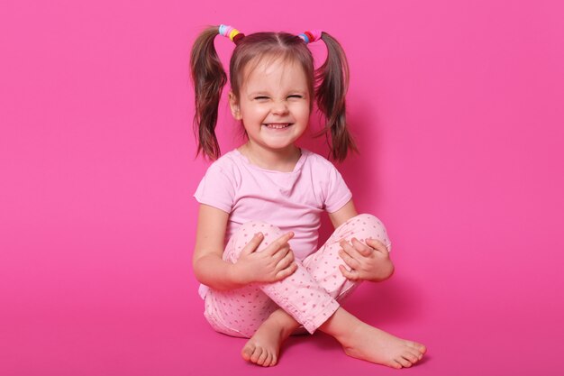 Indoor laughing positive kid sitting on floor, posing isolated on pink, wearing rose t shirt and trousers, having ponytails, being in high spirits. Childhood concept.