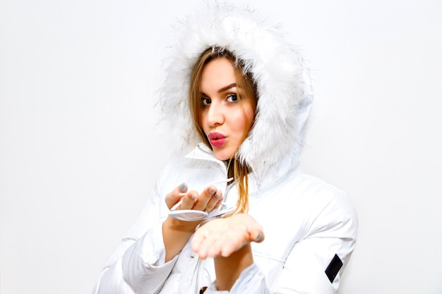 Indoor lifestyle fashion portrait of young blonde woman wearing white winter parka, image with flash, long hairs, natural ,make up, joy, fun.