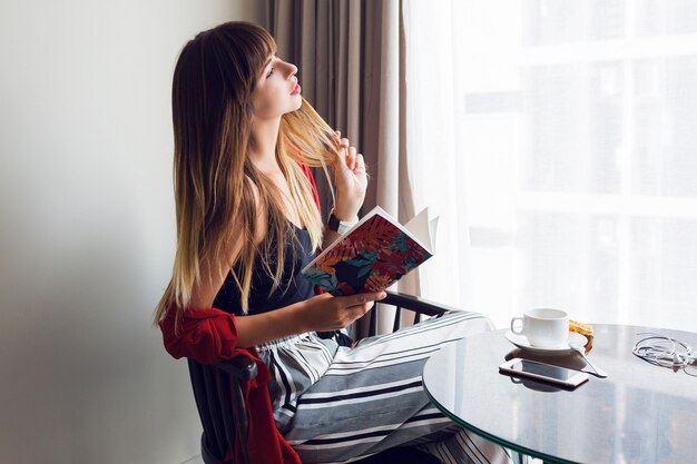 Indoor lifestyle portrait of  pretty brunette woman   reading book  , sitting on chair and drinking coffee in sunny spring morning.  Breakfast time.