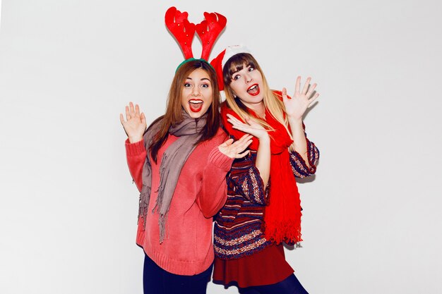 Indoor lifestyle portrait of two best friends.Holiday makeup and cute winter cozy outfit , white wall. Wearing masquerade hats.