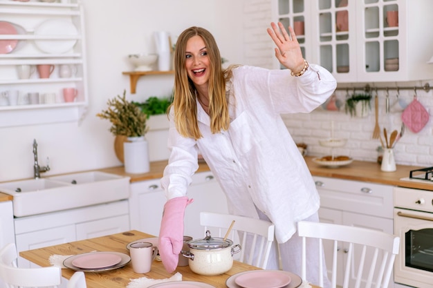 白いリネンのスーツを着た屋内ライフスタイルの肖像画の女性は、彼女のキッチンで食事を準備し、完璧な主婦であり、家で彼女の時間を楽しんでいます。