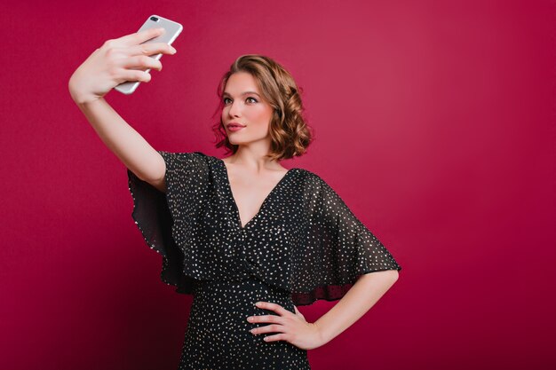 Indoor photo of attractive young lady in vintage dress making selfie on claret background