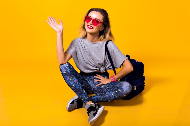 Indoor photo of girl in colorful leggings and trendy shirt sitting on the floor. Studio portrait of stylish young woman in sport shoes posing on yellow background and holding backpack.