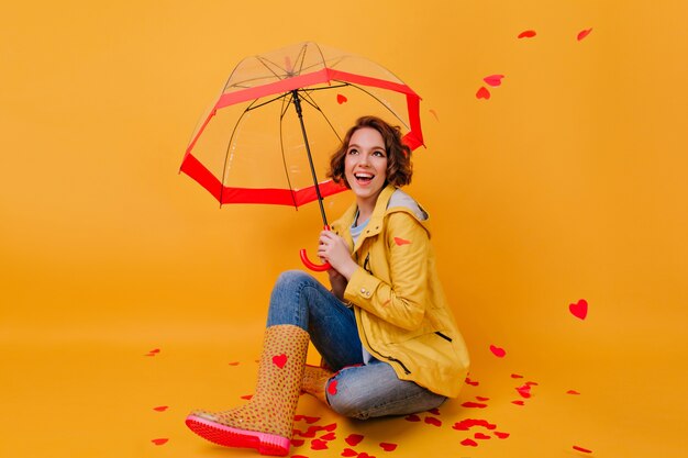 Indoor photo of girl in trendy rubber shoes laughing under umbrella. Studio shot of ecstatic lady fooling around in valentine's day.