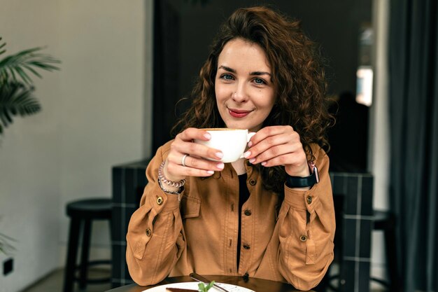 Indoor photo of nice cute girl with wavy hairstyle is dressed brown shirt is holding a cup of coffee and looking at camera with charming smile
