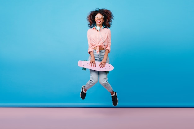 Longboard와 함께 춤을면 핑크 셔츠에 편안한 아프리카 여자의 실내 사진. 여름에 스케이트 보드와 함께 즐기는 갈색 피부와 곱슬 소녀 점프의 초상화.