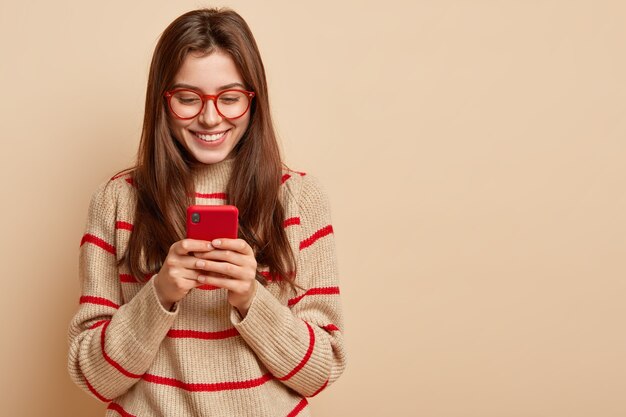 Indoor photo of satisfied teenage girl texts on cellular, reads interesting article online, wears casual outfit, creats new publication on own web page, isolated over brown  wall with free space
