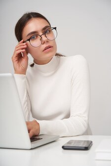 Indoor photo of young pretty brunette woman in glasses looking seriously  while sitting at table over white wall with modern laptop, dressed in formal clothes