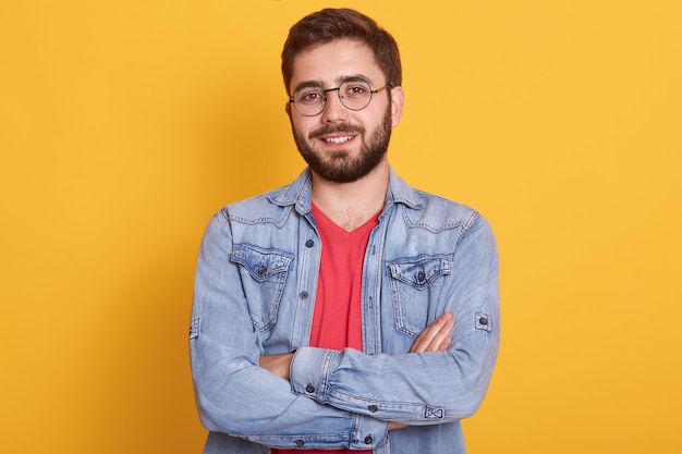 Indoor picture of cheerful handsome young man having folded hands, looking directly  smiling sincerely, wearing casual clothes