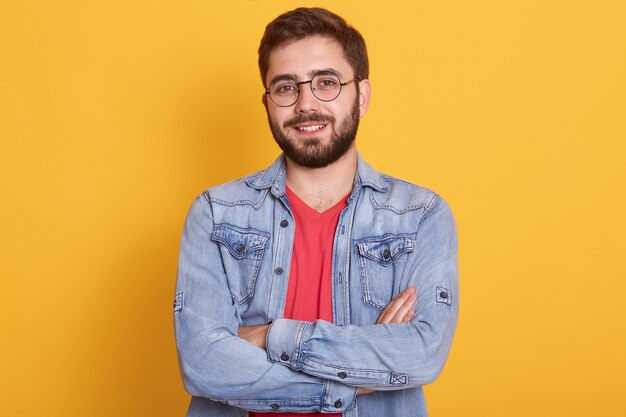 Indoor picture of cheerful handsome young man having folded hands, looking directly  smiling sincerely, wearing casual clothes