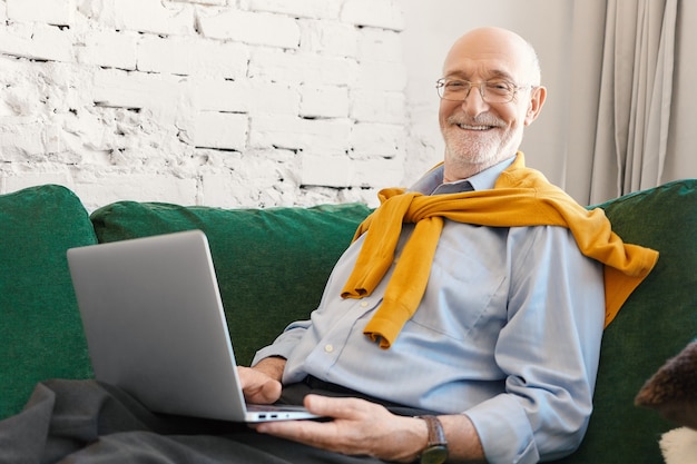 Indoor picture of positive attractive male journalist in spectacles and elegant clothes working on business article for online newspaper or blog, sitting on couch with laptop and smiling at camera