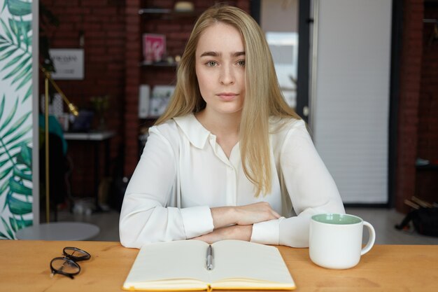 Indoor picture of serious young woman in stylish blouse resting hands on wooden table, having cappuccino during coffee break and writing in copybook
