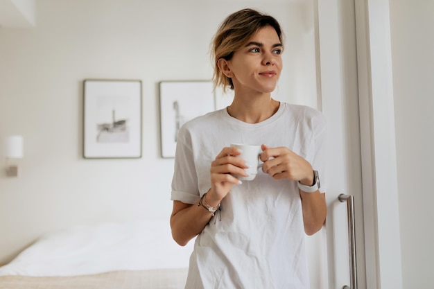 Внутренний портрет молодой женщины в пижаме держит чашку с кофе и смотрит в окно в современной светлой квартире
