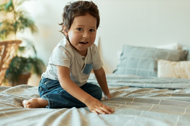 Indoor portrait of adorable infant with dark skin, curly hair and bare feet sitting on bed in t-shirt and jeans