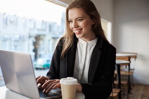 매력적인 유럽 여자 카페에 앉아 커피를 마시고 노트북에 입력, 행복하고 기쁘게 실내 초상화.