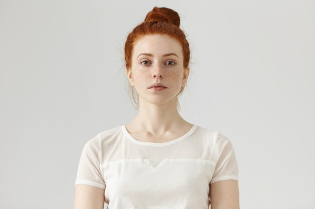 Indoor portrait of attractive young European ginger woman with freckled face and hair bun dressed in white blouse, her look and posture expressing self-confidence