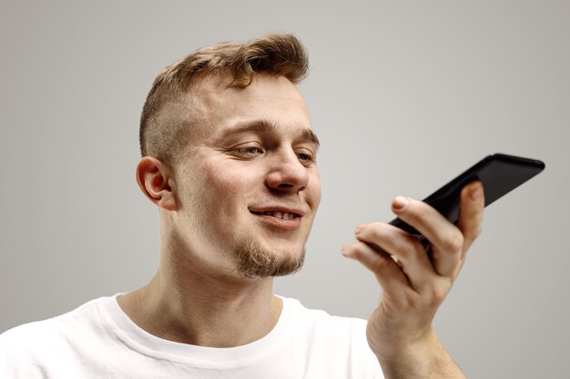 Indoor portrait of attractive young man isolated on gray space, holding smartphone, using voice control, feeling happy and surprised