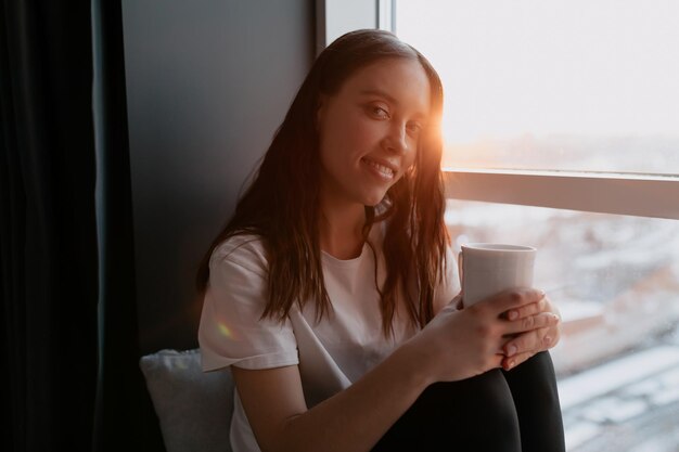 Внутренний портрет удивительно улыбающейся женщины с волнистыми темными волосами и милой улыбкой держит утренний кофе и смотрит в камеру при утреннем солнечном свете Счастливая девушка стоит у окна в солнечном свете
