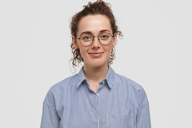 Indoor portrait of beautiful freckled woman with dark curly hair, wears fashionable striped shirt, rejoices day off, isolated over white wall. Curly satisfied woman stands indoor alone