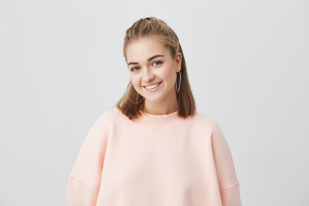 Indoor portrait of beautiful young caucasian woman with straight blonde hair smiling cheerfully, showing her white teeth to camera while feeling happy and carefree on her days-off