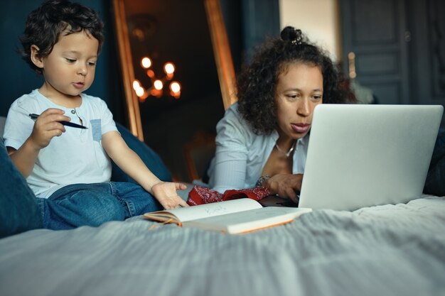 그녀의 귀여운 작은 아들이 연필을 들고있는 동안 노트북과 침대에 누워 집에서 원격으로 작업하는 아름 다운 젊은 여성 프리랜서의 실내 초상화