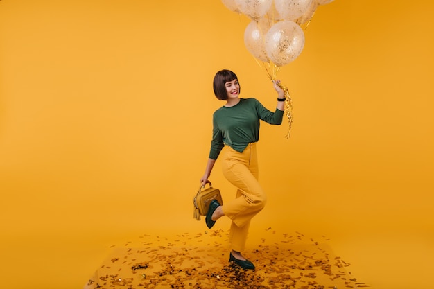 Indoor portrait of birthday girl posing on one leg and smiling. Cute white woman in yellow pants dancing.