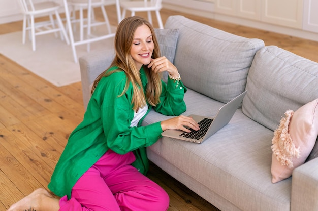 明るいシャツとズボンを身に着けているブロンドのフリーランサーのブロンドの女性の屋内の肖像画は、リビングルームのホームオフィスのコンセプトで彼女のラップトップに取り組んでいます