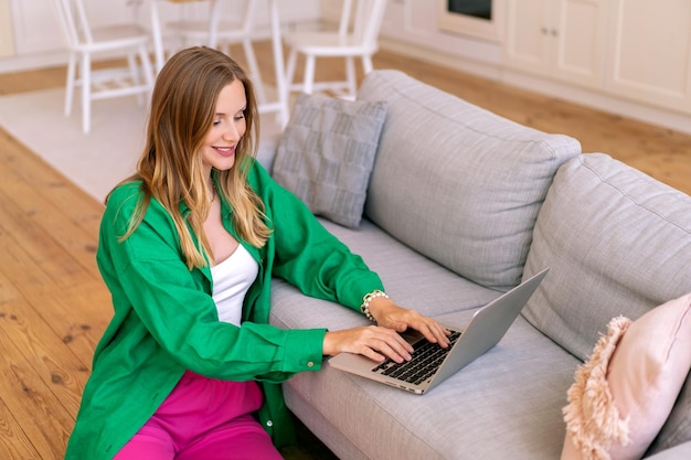 금발 프리랜서 금발 여성의 실내 초상화는 밝은 셔츠와 바지를 입고 거실 홈 오피스 컨셉에서 노트북 작업을 하고 있습니다.