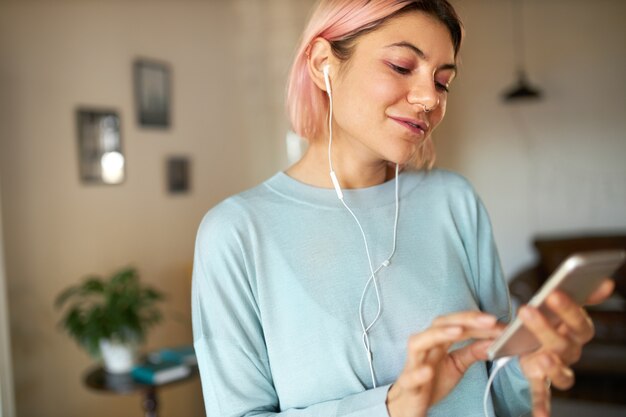 Indoor portrait of charming beautiful teenage girl with pink hair and eyeshadows relaxing at home using earphones