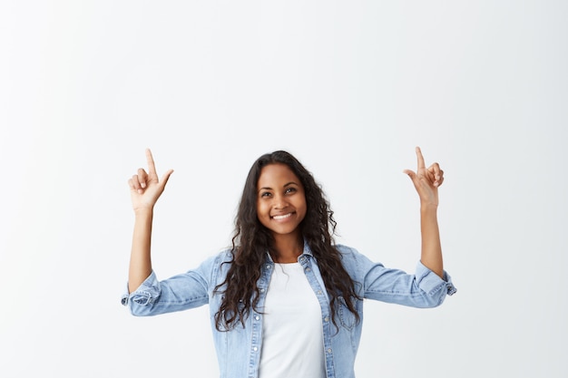 Indoor portrait of cheerful attractive afro-american girl casually dressed with long wavy hair pointing her index fingers up, indicating something interesting, having happy excited look.