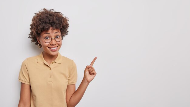 Indoor portrait of cheerful good looking woman points index finger at upper right corner suggests to place your advertisement here smiles happily wears spectacles and beige t shirt poses indoor