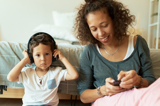 Indoor Portrait of cheerful young Hispanic female holding mobile phone playing music tracks for her adorable baby son who listening to songs via wireless headphones