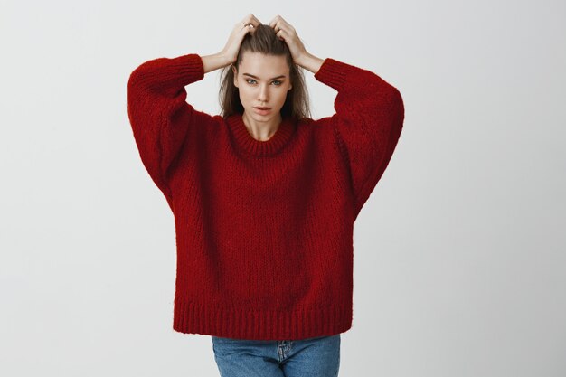 Indoor portrait of confident attractive caucasian businesswoman in stylish red sweater, touching hair and looking focused , posing sensually  for advertisement
