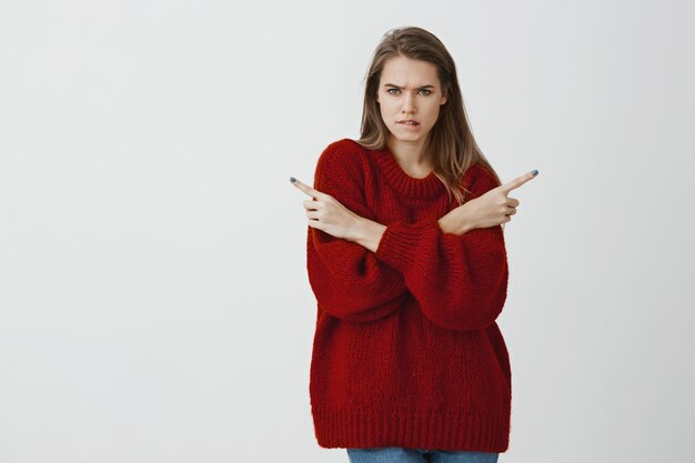 Indoor portrait of confused troubled attractive woman in stylish loose sweater, biting lip, frowning and pointing in different directions, being questioned and displeased, having problem to choose