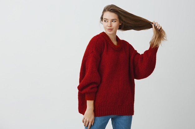 緩い赤いセーターを着て好奇心が強いロマンチックな白人女性の屋内ポートレート、美しい健康な髪を手に持って、情熱的なリラックスした気分で左を見て、立っています。