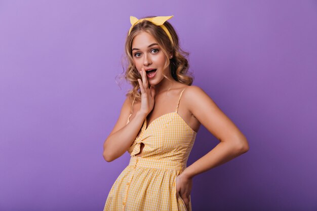 Indoor portrait of curious tanned woman in yellow dress. Adorable curly white girl looking to camera.