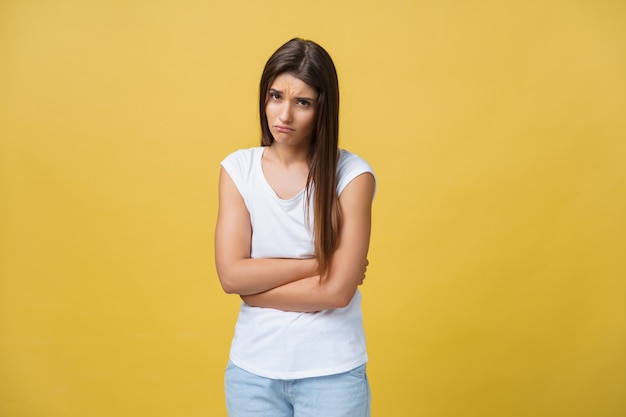 Indoor portrait of cute girl standing with crossed hands on belly, feeling awkward or suffering from pain while looking aside, standing against yellow background. Woman has stomachache.