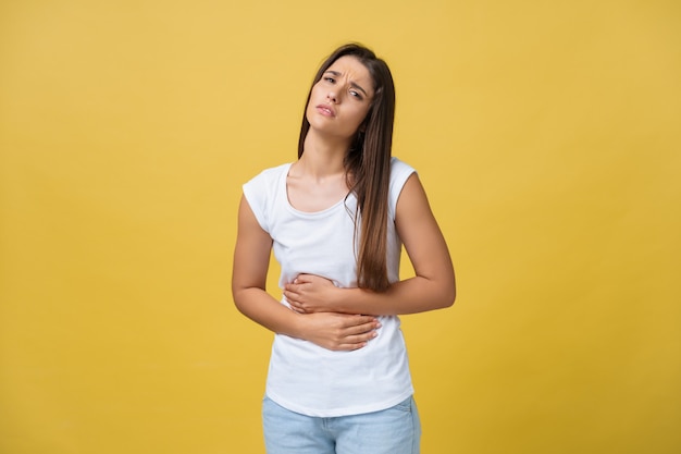 Indoor portrait of cute girl standing with crossed hands on belly, feeling awkward or suffering from pain while looking aside, standing against yellow background. Woman has stomachache