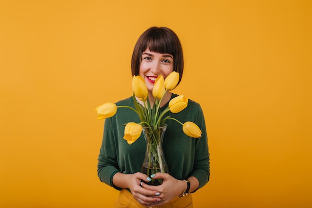 Foto gratuita ritratto dell'interno della ragazza dai capelli scuri in maglione verde che tiene un vaso di bellissimi fiori. signora estatica con i capelli corti in piedi con i tulipani.