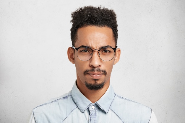 Indoor portrait of displeased discontent upset male model with trendy hairdo, mustache, beard, wears round glasses