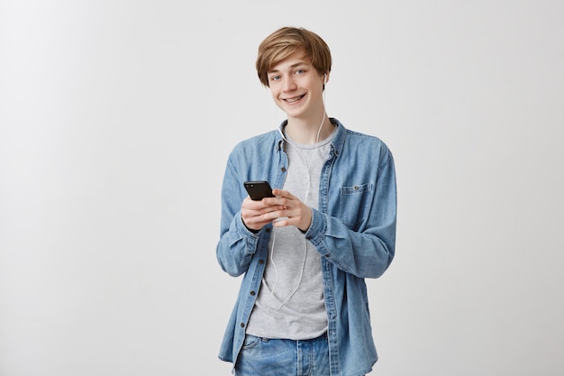 Indoor portrait of european fair-haired guy with blue eyes in denim shirt holding mobile phone messaging with friends telling them funny stories, has pleasant smile standing 