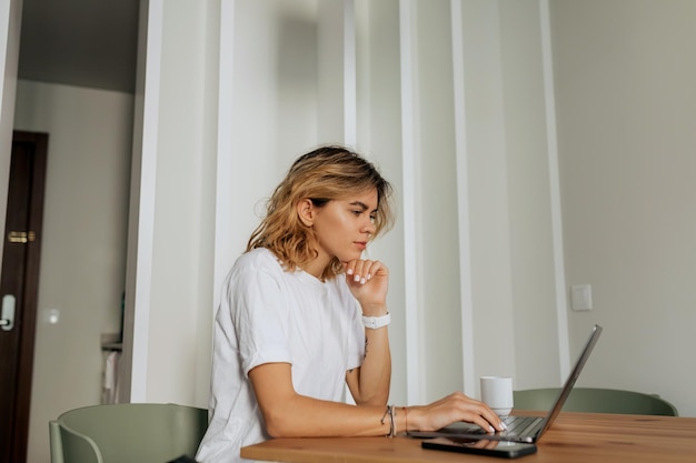 Indoor portrait of European woman with wavy light hair wearing white tshirt is working with laptop at home Working woman at home office in warm sunny day