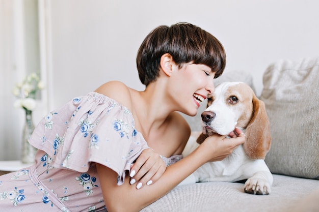 光沢のある濃い茶色の髪の傷を持つ興奮した若い女性の屋内の肖像画は笑顔でビーグル犬を喜ばせた