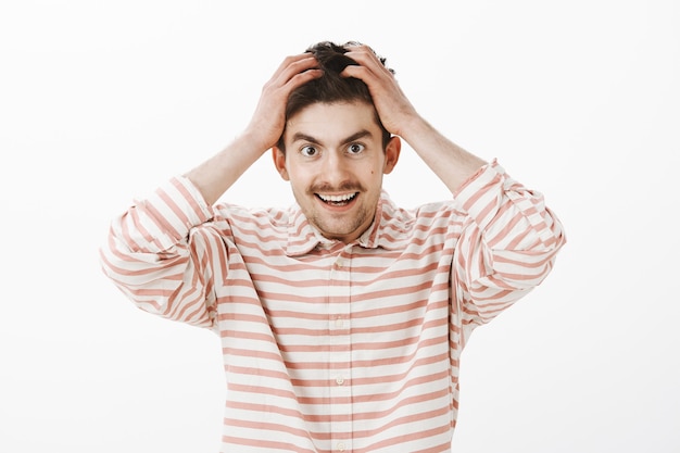 Indoor portrait of fascinated lucky male model with moustache, holding hands on hair and smiling happily, doubting his own luck, standing against gray wall overwhelmed with great emotions