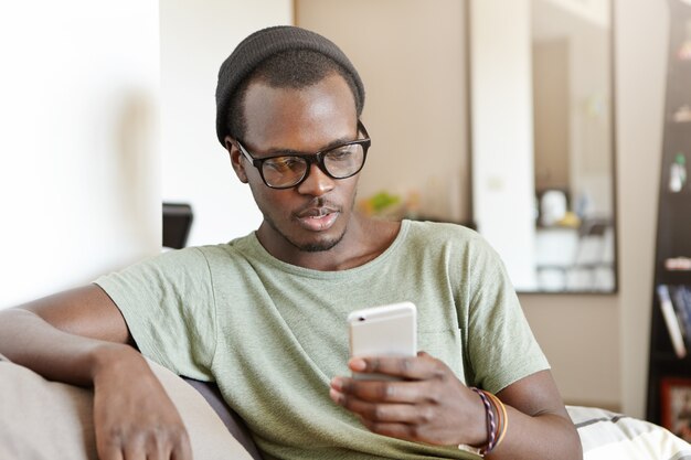 Крытый портрет модного молодого афроамериканского человека, отдыхающего дома, сидящего на диване с сенсорным мобильным телефоном, с помощью онлайн-приложения