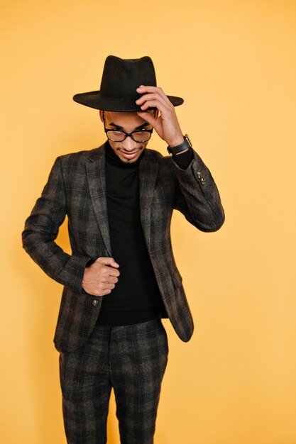 Indoor portrait of glad african male model playfully posing on yellow wall. Confident black young man in jacket touching his hat during photoshoot.