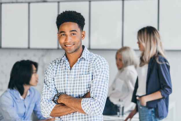 免费室内的肖像照片高兴非洲学生在蓝色衬衫站在双手交叉,而他的大学朋友说在他身边。幸福的黑人把时间花在办公室与同事。