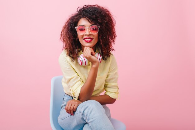 Indoor portrait of glamorous black woman with inspired smile sitting in chair and gently touching her chin. Laughing mulatto female model in trendy jeans and yellow jacket posing.